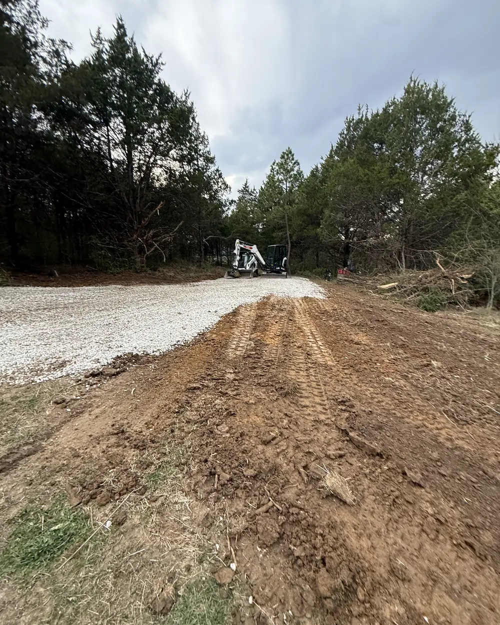 A strip of ground cleared of brush and cut to grade beside a new gravel drive, with the cleared timber stacked along the treeline