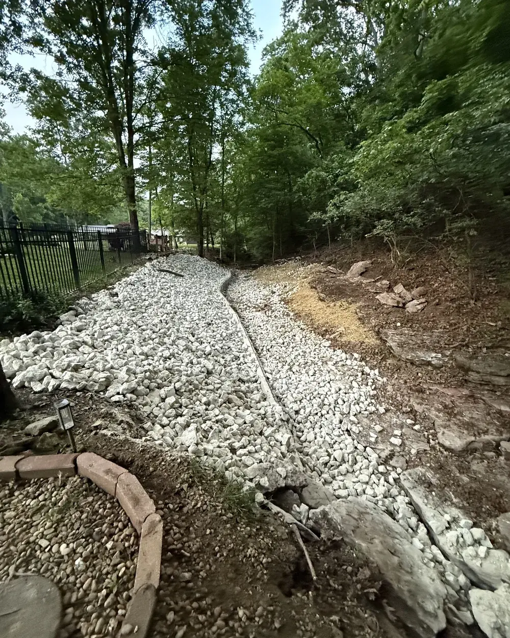 A finished drainage run of white rock cut along a wooded slope behind a black iron fence, with the regraded bank seeded beside it