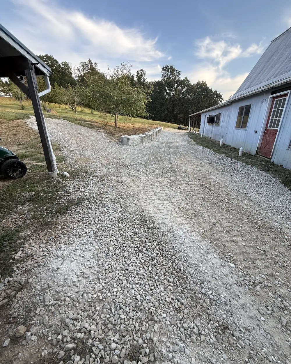 A new gravel driveway laid between a white timber barn and a carport, with a low block wall retaining the bank beside it