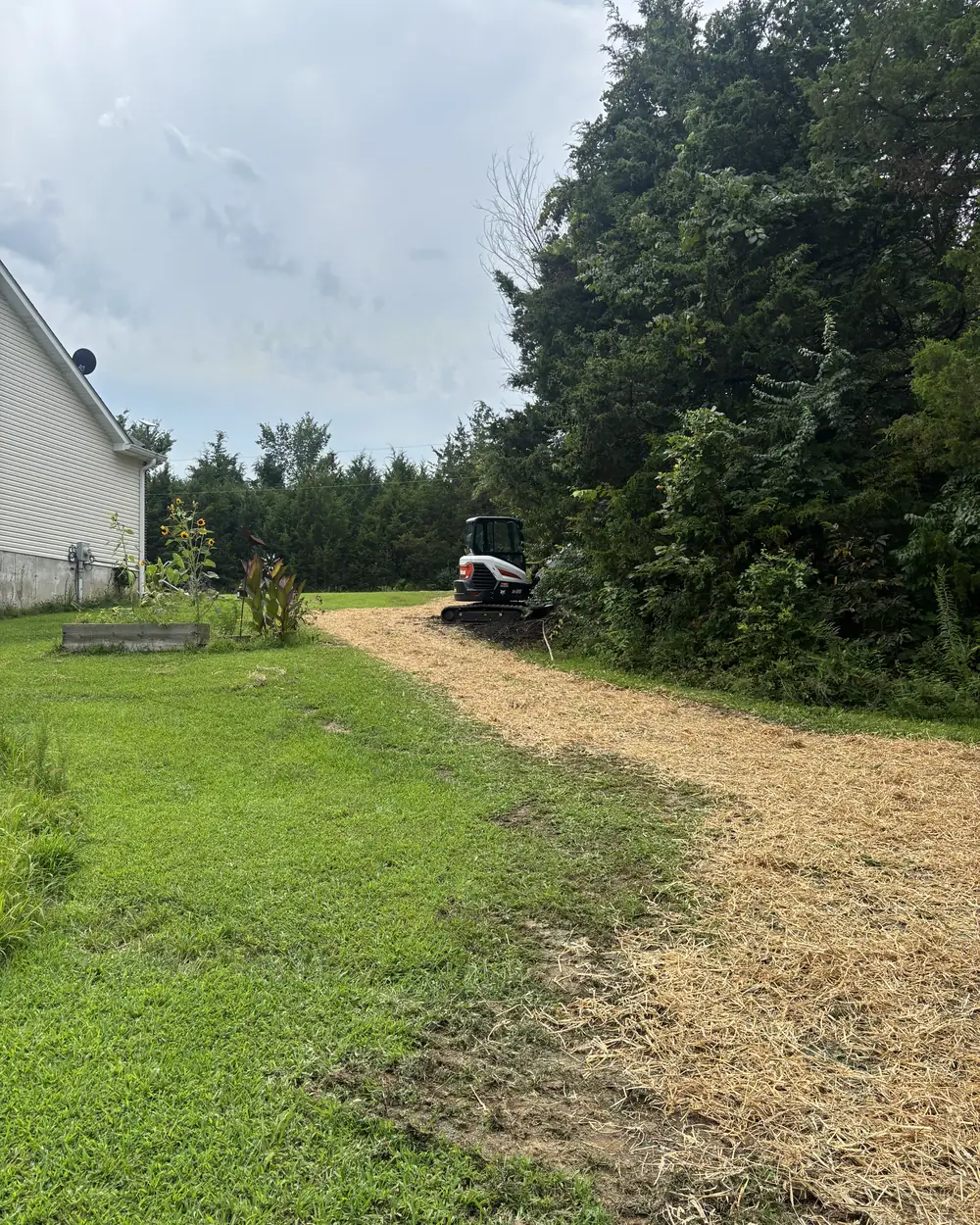 A graded strip of ground beside a house seeded and covered in straw, with a compact excavator parked at the far end of it