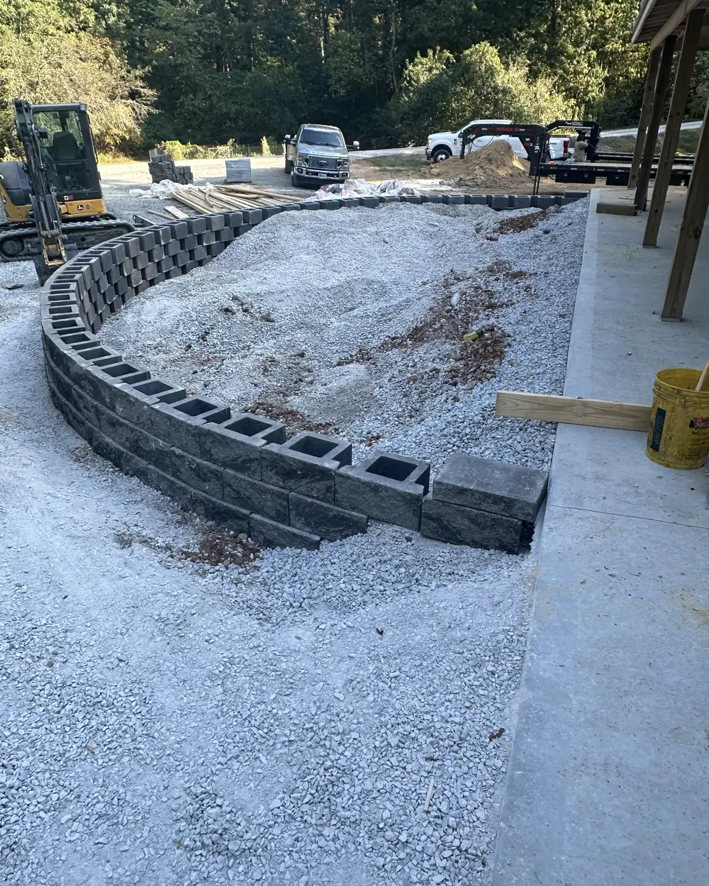 A curved retaining wall part-built, its block courses set in a sweep with a compact excavator behind it and gravel backfill inside the curve
