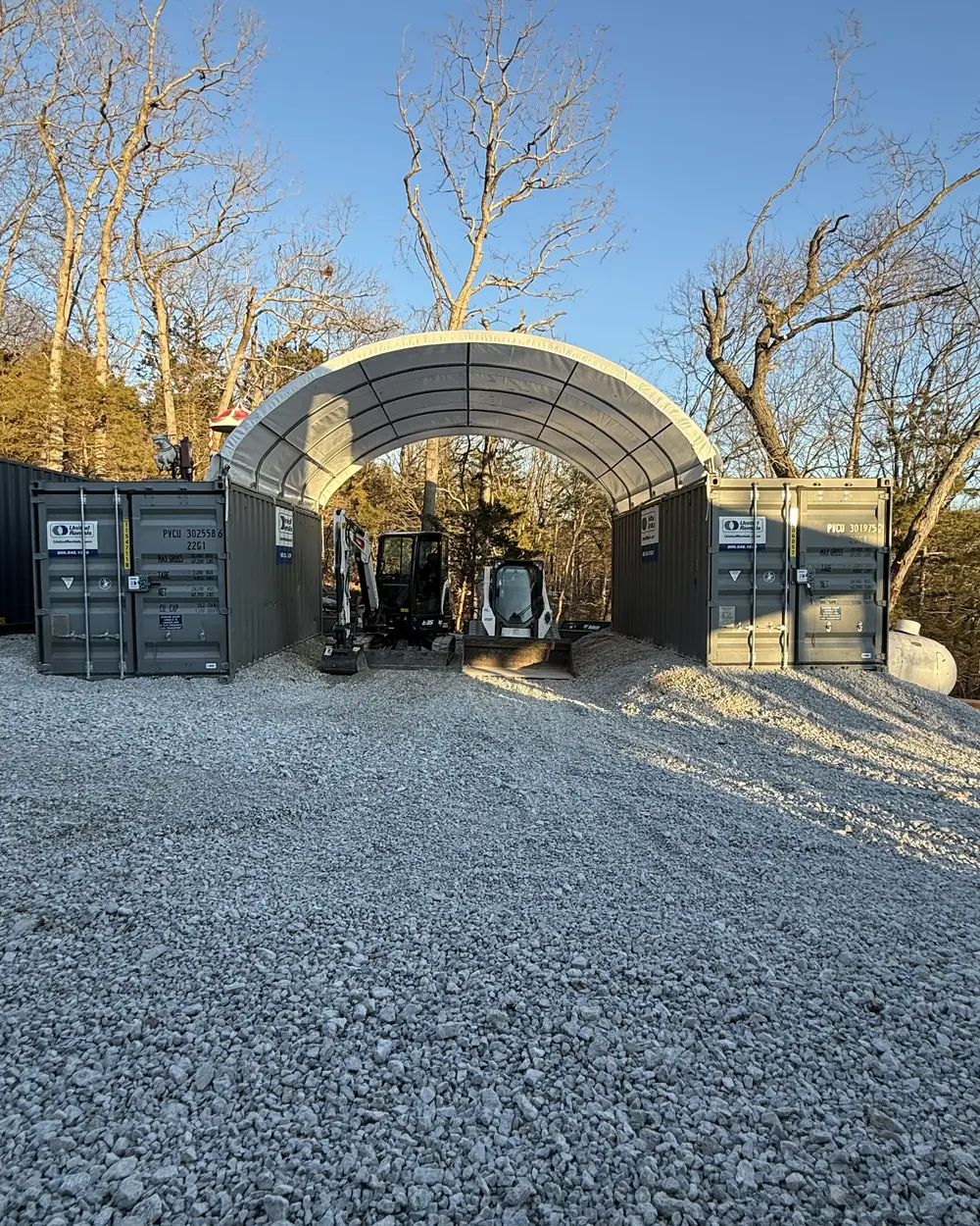 Two compact machines parked under an arched shelter between shipping containers in the company's own gravel yard